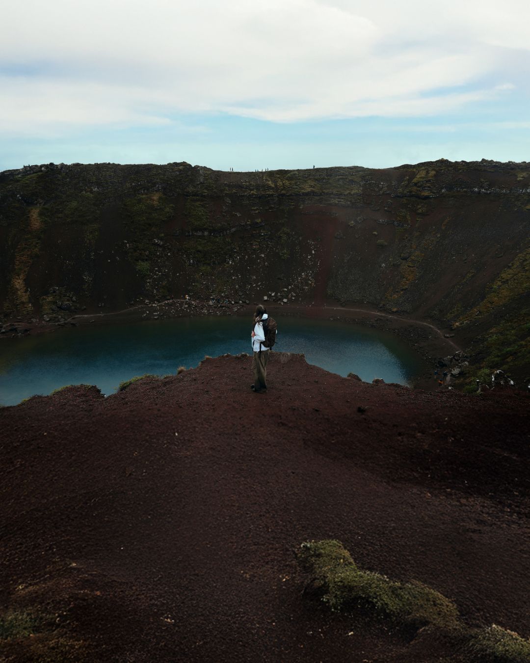 A person stands on a dark volcanic cliff overlooking a crater lake with steep dark walls and a cloudy sky.