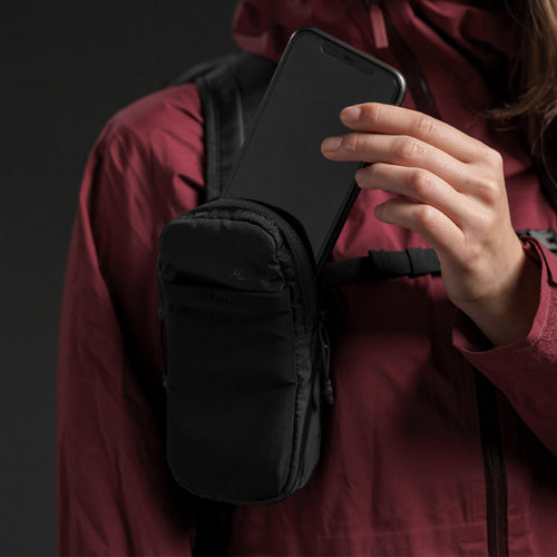 Woman in burgundy jacket, placing phone inside of speed stash, attached to her backpack strap